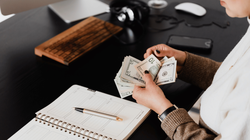 Kobieta w beżowym swetrze siedzi przy ciemnym biurku, licząc dolary. Obok niej na biurku leżą notatnik, długopis oraz inne przedmioty biurowe, takie jak klawiatura i mysz komputerowa, co sugeruje, że pracuje z domu.
