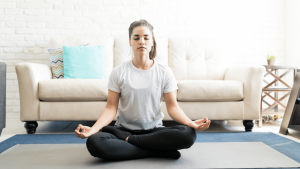 Młoda kobieta w białej koszulce i czarnych leginsach medytuje w pozycji siedzącej na niebieskiej macie do jogi. Znajduje się w jasnym pokoju z białymi ścianami, a w tle widać sofę i dekoracyjne poduszki.