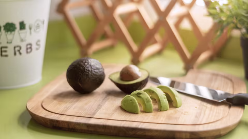 Zdjęcie przedstawia deskę do krojenia z kawałkami awokado. Na desce znajduje się również połówka awokado z pestką oraz cały owoc. Obok deski leży nóż kuchenny. W tle widoczny jest biały pojemnik z napisem "HERBS" oraz rośliny, co sugeruje przytulną kuchenną atmosferę.