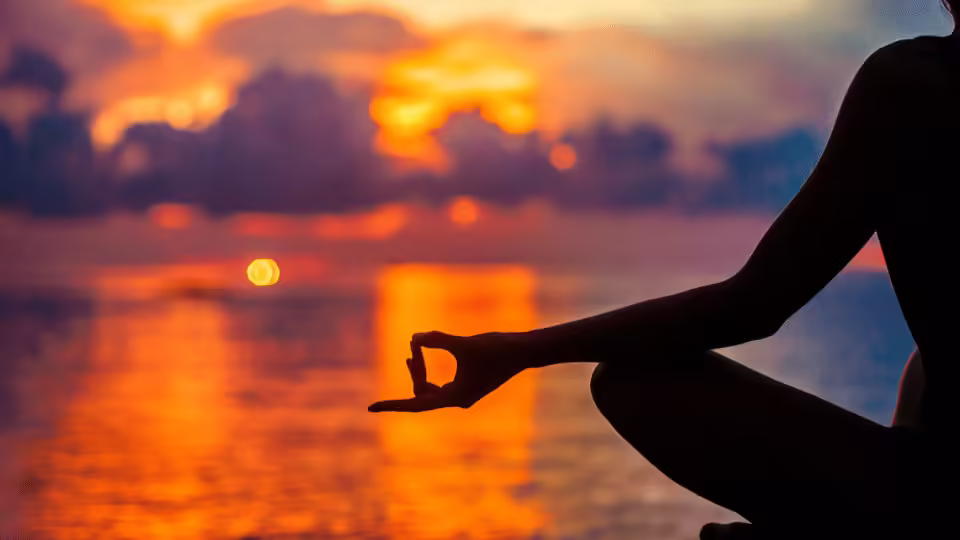 Ręka osoby medytującej o zachodzie słońca, tworząca gest mudry, na tle spokojnego jeziora i kolorowego nieba, symbolizując harmonię i wewnętrzny spokój podczas praktyki afirmacji.