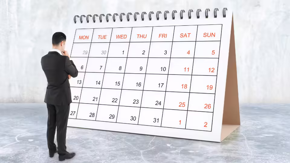 Mężczyzna w garniturze patrzący na duży kalendarz, zastanawiający się nad planowaniem lub wyznaczaniem terminów, co może symbolizować refleksję nad czasem potrzebnym na osiągnięcie celów lub realizację afirmacji.