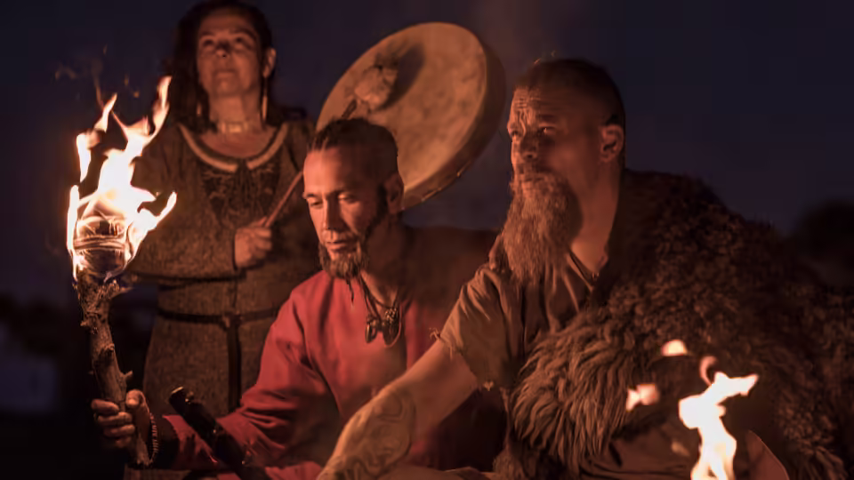 Trzech postaci w nordyckich strojach tradycyjnych bierze udział w rytualnym obrzędzie przy ognisku. Jedna z nich trzyma płonącą pochodnię, a w tle kobieta gra na bębnie. Scena jest oświetlona ciepłym blaskiem ognia, co podkreśla atmosferę tajemniczości i duchowości, charakterystyczną dla starożytnych ceremonii Wikingów.