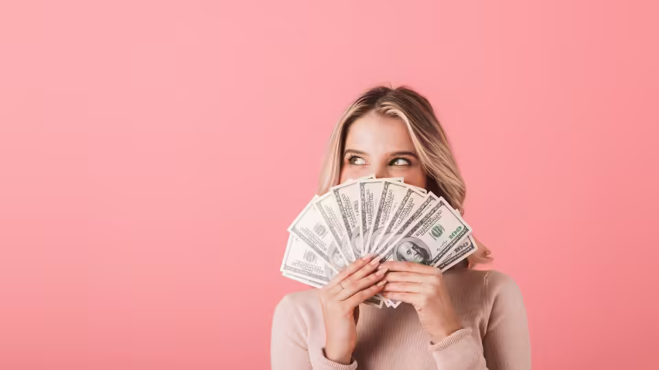 Kobieta trzymająca przed sobą wachlarz banknotów dolarowych na tle w kolorze różowym, symbolizująca pomyślność finansową i obfitość.