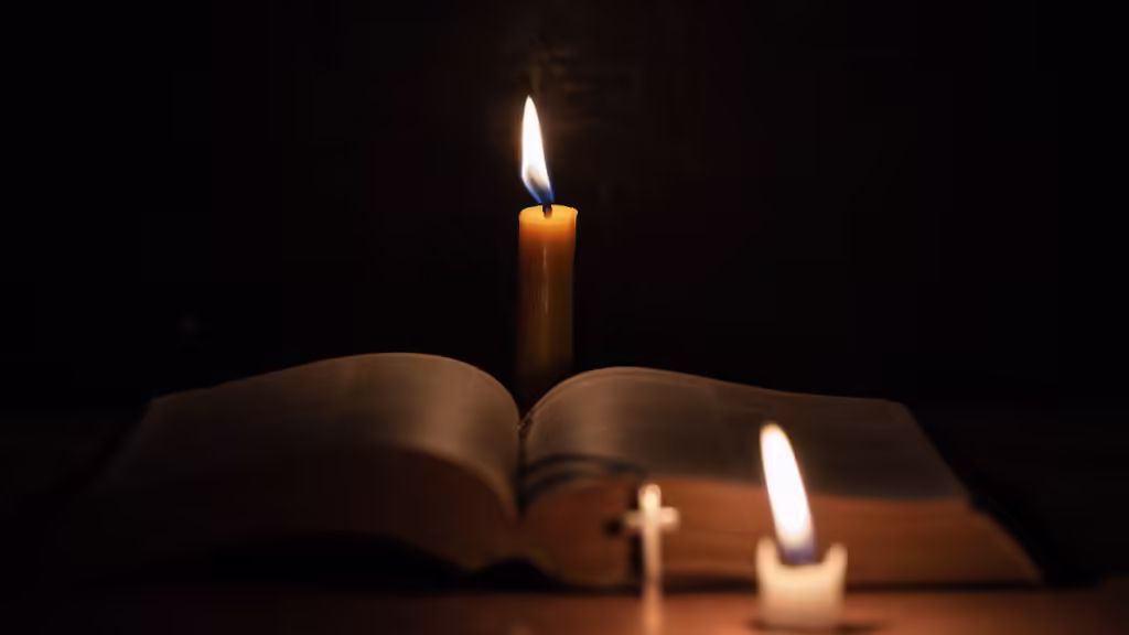 Otwarta księga z krzyżem i zapalonymi świecami w ciemnym otoczeniu, symbolizująca duchowość i modlitwę.