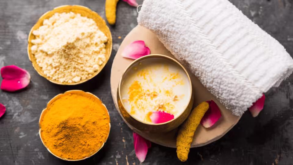 Zestaw do pielęgnacji skóry z naturalnymi składnikami: miseczka z mieszanką na bazie kurkumy, mąki oraz napój z płatkami róż, obok ręcznik i płatki kwiatów na kamiennej powierzchni.