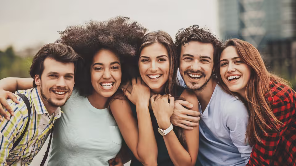 Grupa przyjaciół w różnym wieku, szeroko uśmiechniętych i obejmujących się, emanujących radością i pozytywną energią.