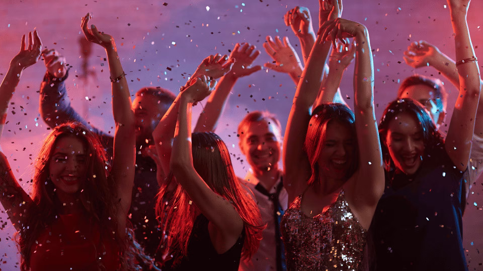 Grupa ludzi świętuje Sylwestra, ubrana w kolorowe stroje – czerwień, złoto i błyszczące tkaniny, symbolizujące energię, luksus i szczęście, w otoczeniu konfetti i radosnej atmosfery.