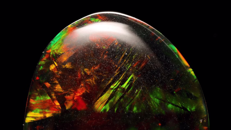 Zbliżenie na czarny opal z intensywnymi, ognistymi refleksami zieleni, czerwieni i pomarańczu na tle głębokiej czerni, podkreślające jego mistyczny urok.