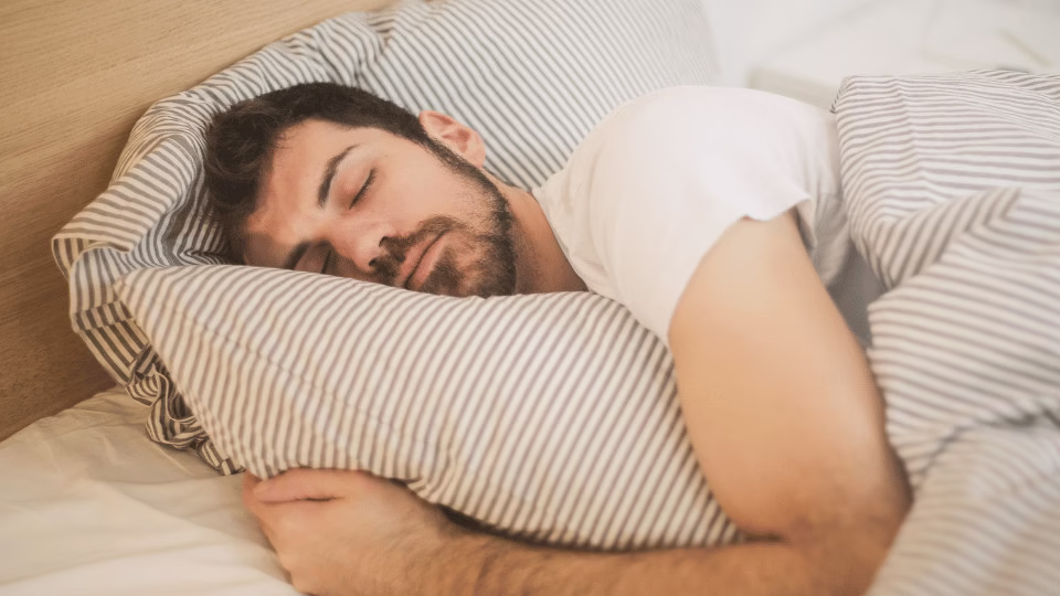 Mężczyzna w białej koszulce śpi spokojnie na łóżku, obejmując poduszkę w biało-szare paski, co symbolizuje regenerujący sen w relaksującym otoczeniu.
