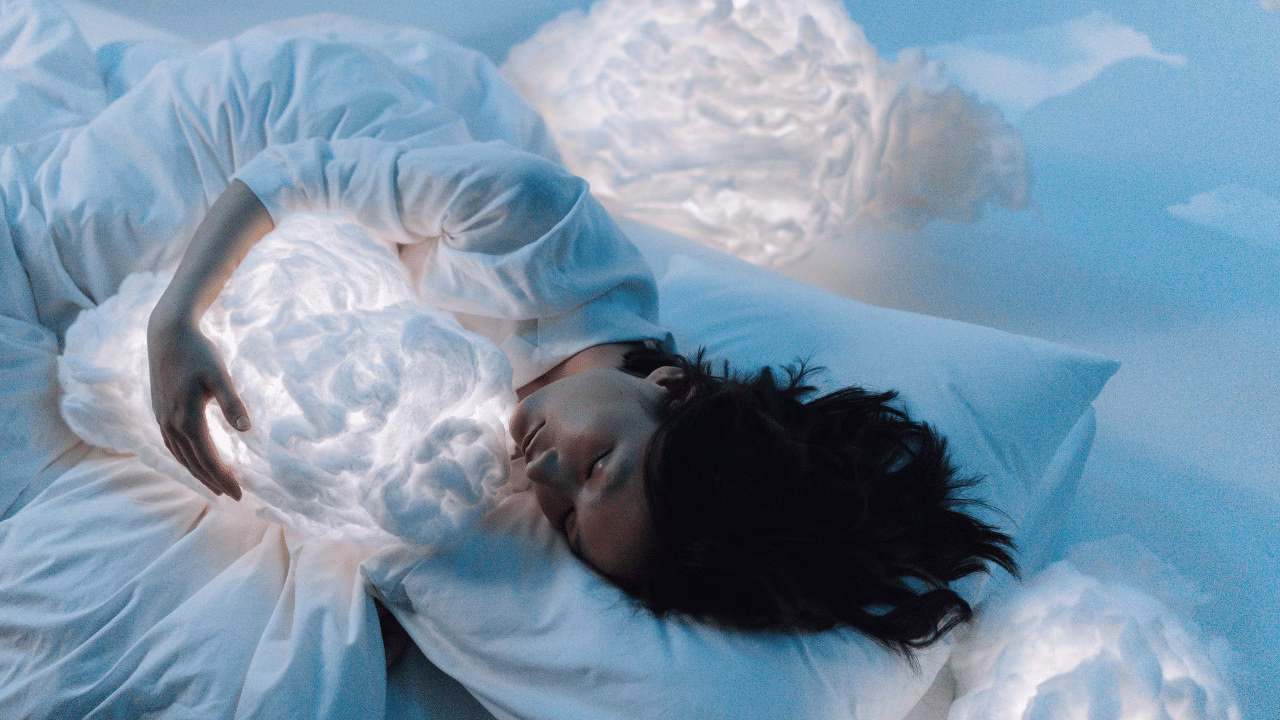 Osoba śpiąca w białej pościeli, przytulająca świecącą poduszkę w kształcie chmury – symbol spokojnego snu i relaksu