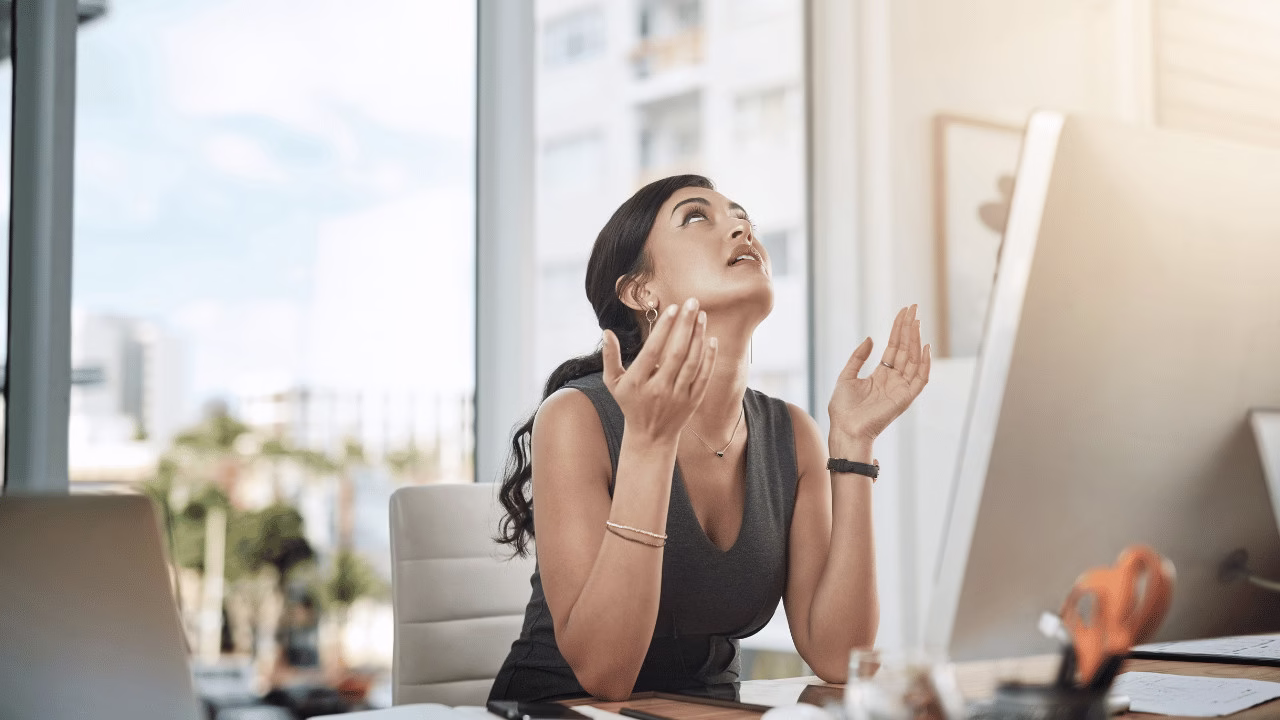 Kobieta siedzi przy biurku w nowoczesnym biurze, patrzy w górę z frustracją i unosi ręce w geście bezsilności.