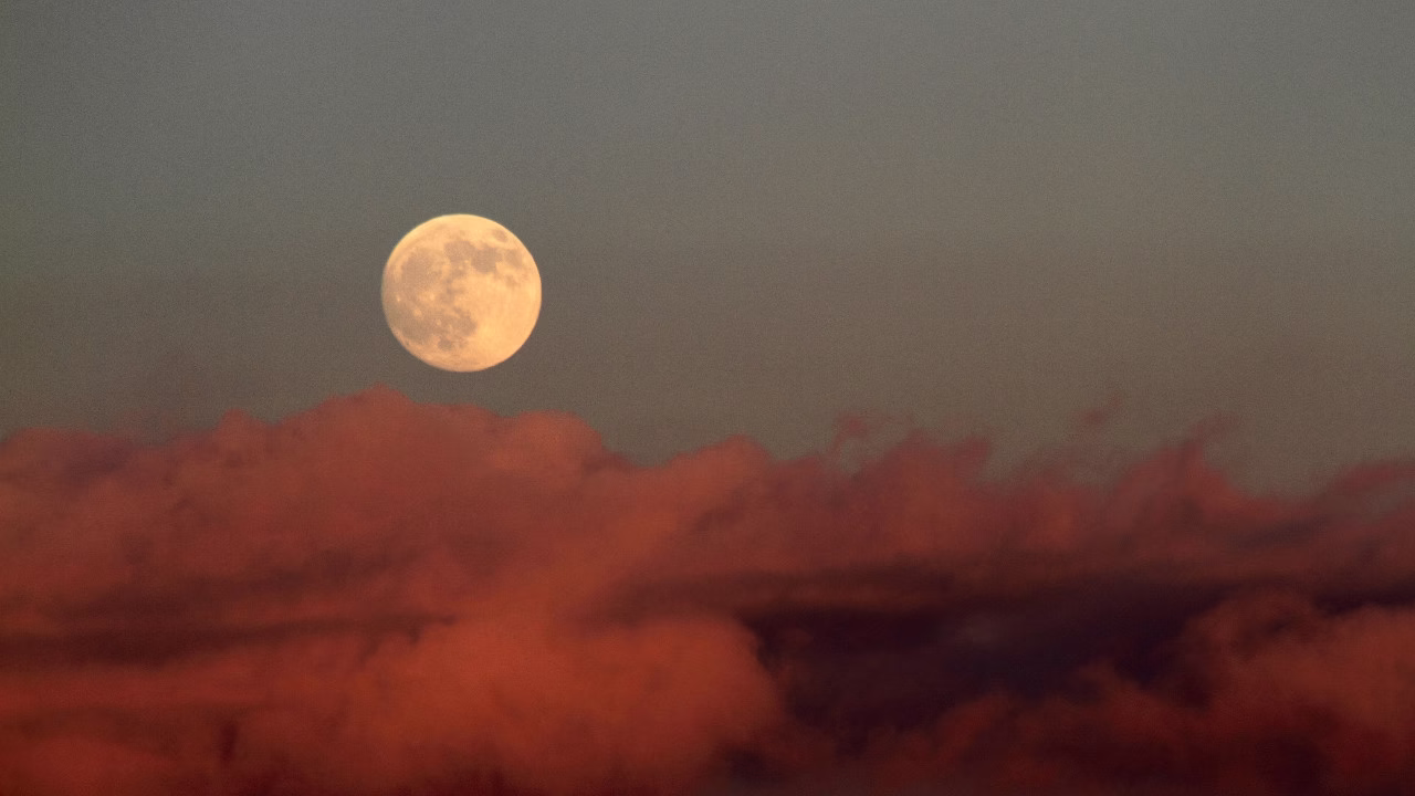 Pełnia Księżyca unosząca się nad pomarańczowo-czerwonymi chmurami o zmierzchu.