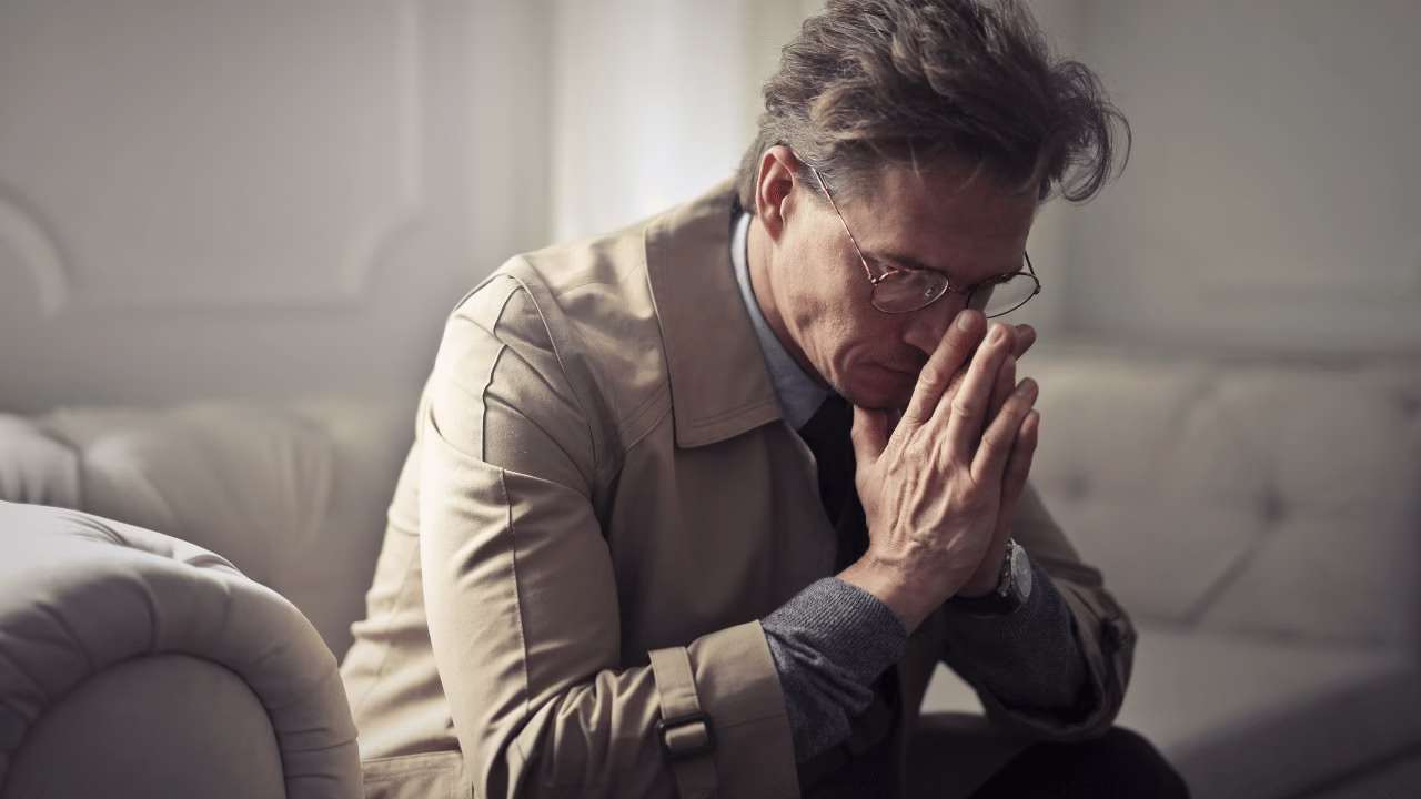 Zamyślony mężczyzna w okularach siedzący na sofie z dłońmi przy twarzy, wyrażający smutek i refleksję