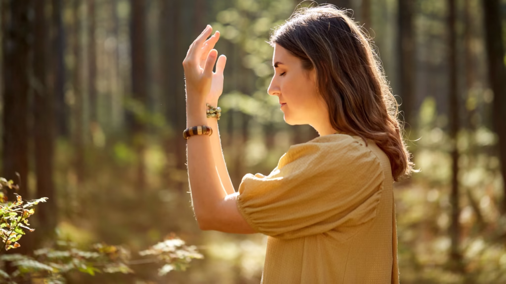 Młoda kobieta w żółtej sukience stojąca w lesie, z zamkniętymi oczami i uniesioną dłonią w medytacyjnym geście.