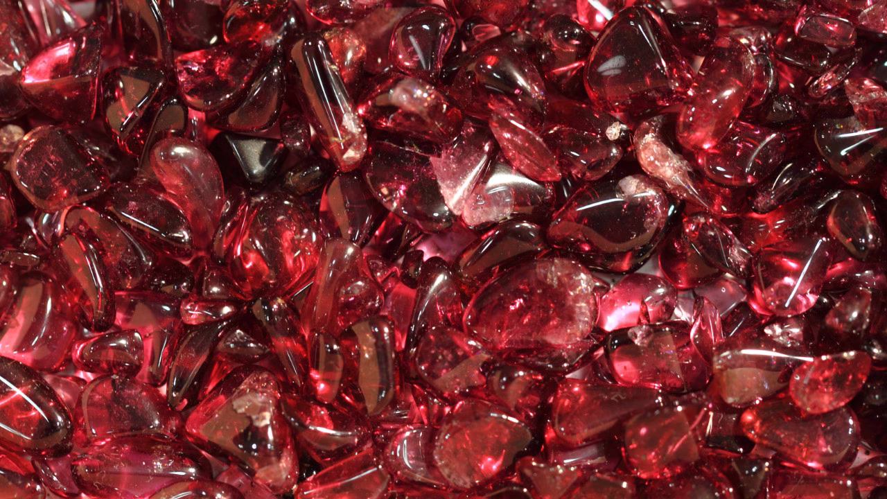 Zbliżenie na skupisko drobnych, gładkich kamieni w intensywnym, przezroczystym czerwonym kolorze. Kamienie mają różne kształty i połyskują w świetle, tworząc efekt błyszczącej, czerwonej mozaiki.
