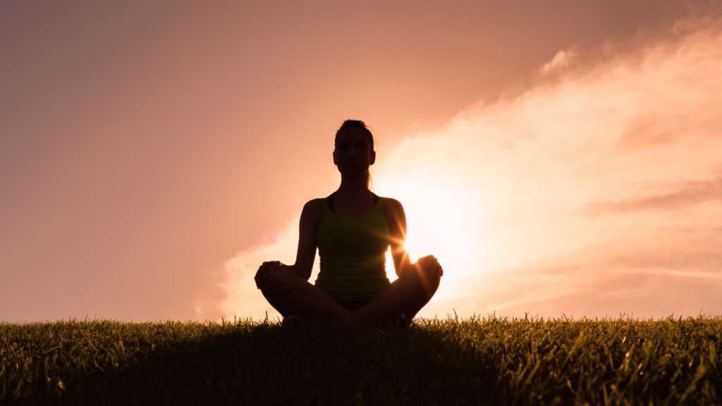 Osoba medytująca w pozycji lotosu na trawie, sylwetka na tle zachodzącego słońca i nieba w ciepłych odcieniach.