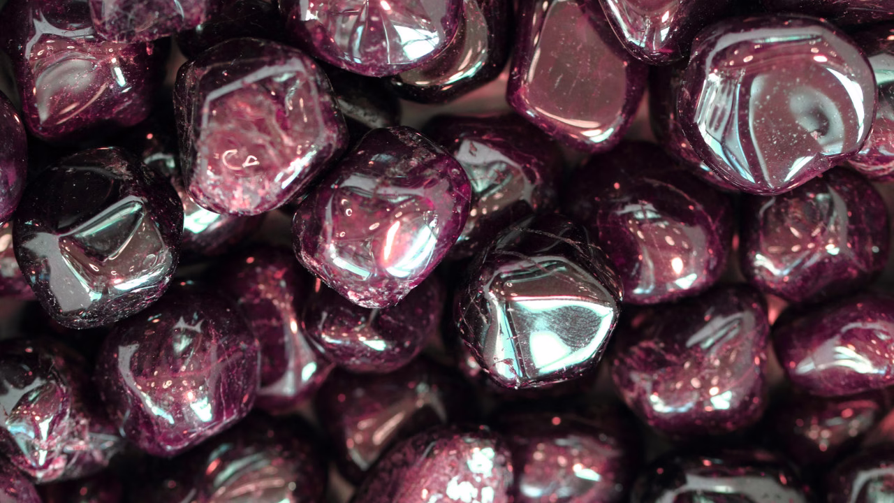 Zbliżenie na skupisko gładkich, wypolerowanych kamieni w ciemnofioletowym kolorze z połyskiem. Kamienie mają zaokrąglone kształty i odbijają światło, nadając powierzchni efekt szklisto-metaliczny.