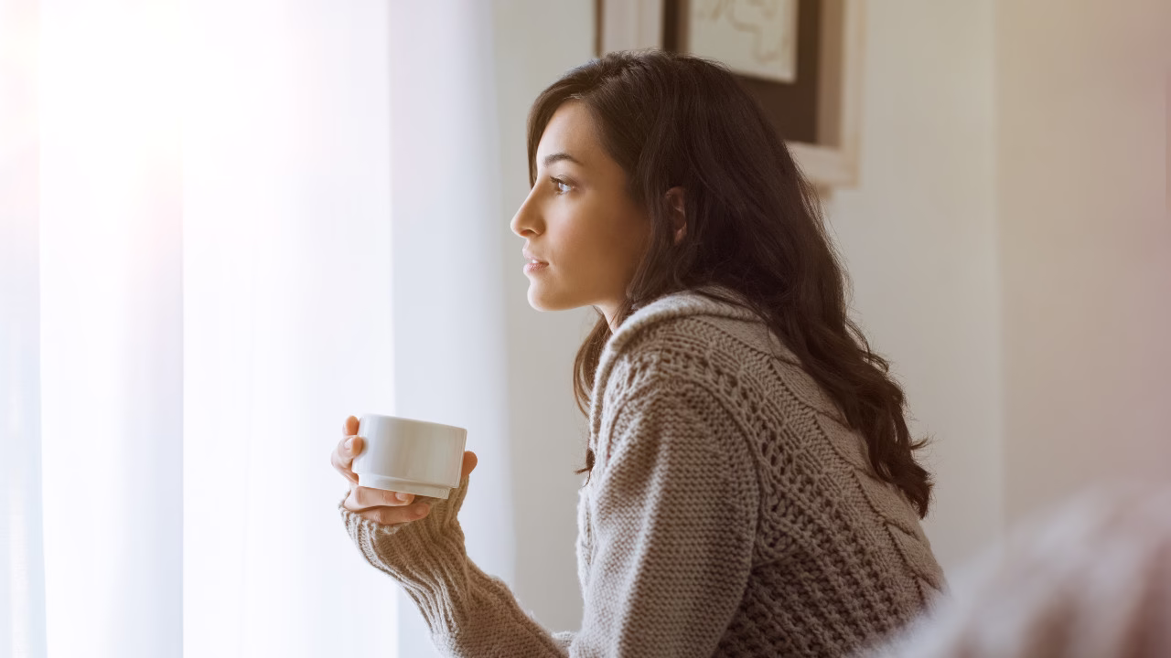 Kobieta w ciepłym swetrze siedzi przy oknie, trzymając kubek z napojem i patrząc zamyślona w stronę światła.
