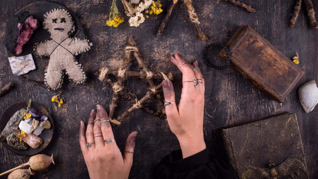 Widok z góry na rytualne przedmioty na drewnianym stole: lalka z tkaniny, pentagram z gałązek, suszone zioła, minerały i dłonie z pierścieniami układające symbole.