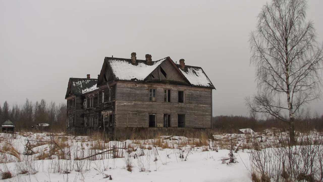 Stary, drewniany, opuszczony budynek w zimowym krajobrazie z zaśnieżonym dachem i wyschniętą roślinnością wokół.