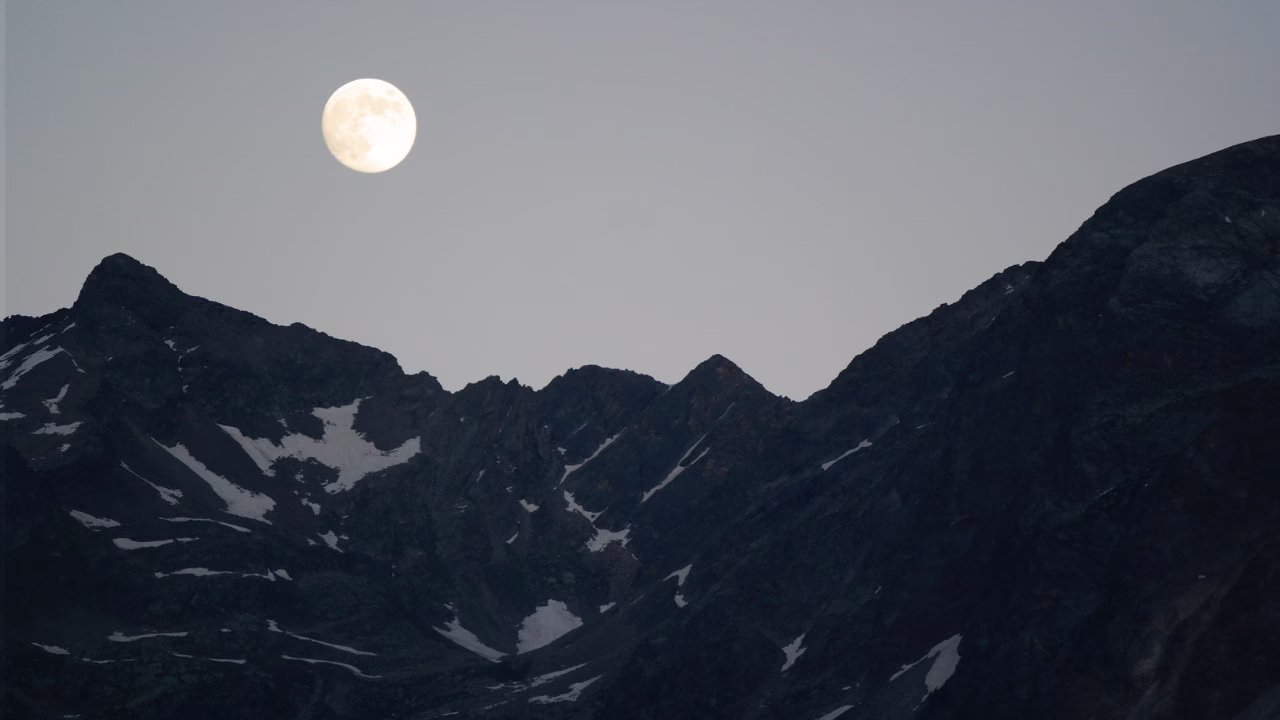 Jasny Księżyc w pełni unoszący się nad ciemnym górskim krajobrazem o zmierzchu, z widocznymi ośnieżonymi fragmentami skał.