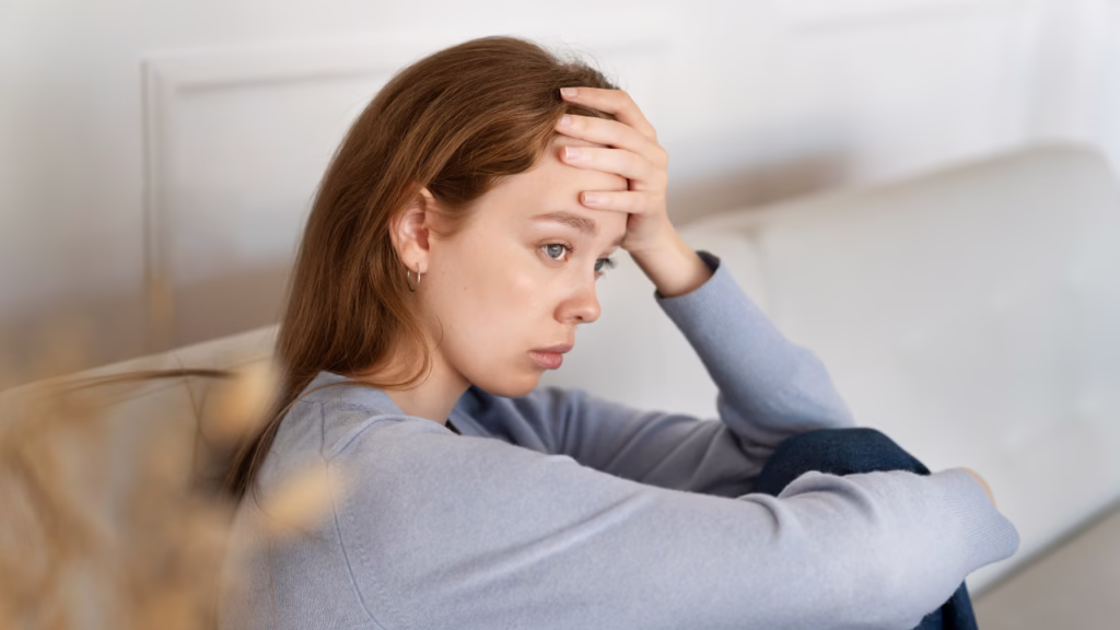 Młoda kobieta siedząca na kanapie, oparta o kolano, z zatroskaną i przygnębioną miną.