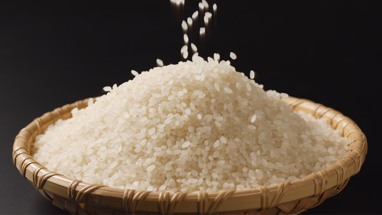 Zbliżenie na wiklinową miskę wypełnioną białym ryżem, nad którą spadają pojedyncze ziarenka na czarnym tle.