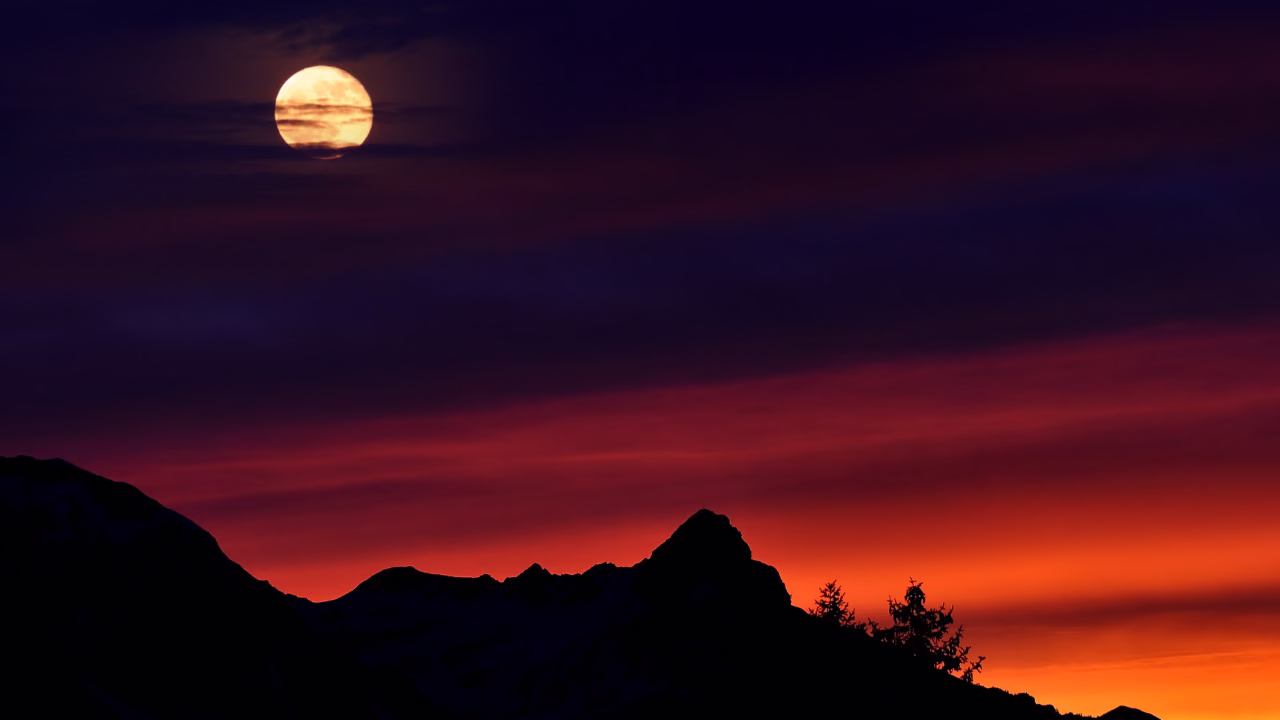 Księżyc widoczny nisko nad horyzontem podczas zachodu słońca, na tle czerwono-pomarańczowego nieba i ciemnych sylwetek gór.