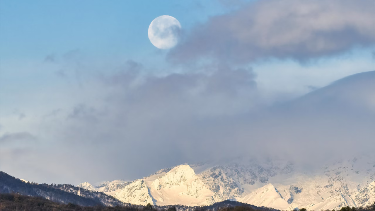 Pełnia Księżyca nad ośnieżonymi górami częściowo przesłonięta chmurami na zimowym niebie