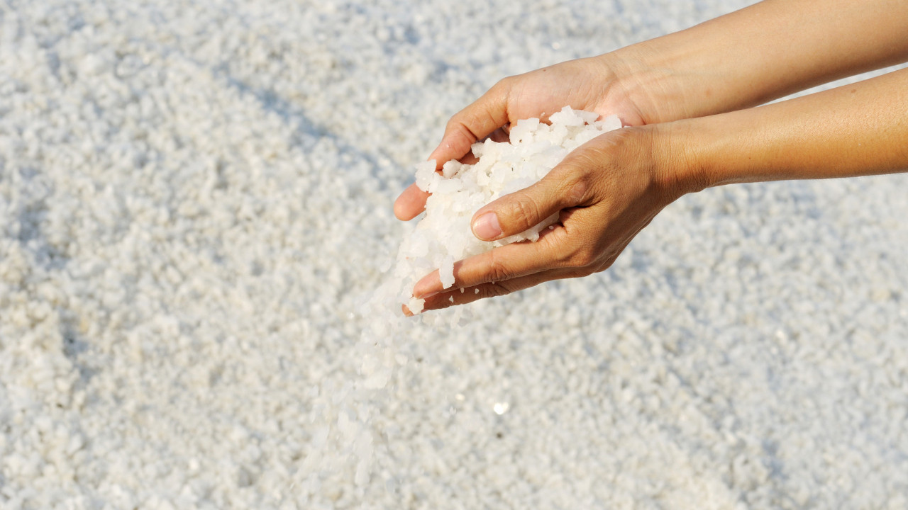 Dłonie trzymające i przesypujące gruboziarnistą białą sól na tle solnego podłoża w naturalnym świetle.