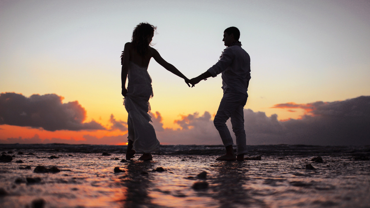 Para trzymająca się za ręce spaceruje po plaży o zachodzie słońca, romantyczny moment zakochanych.