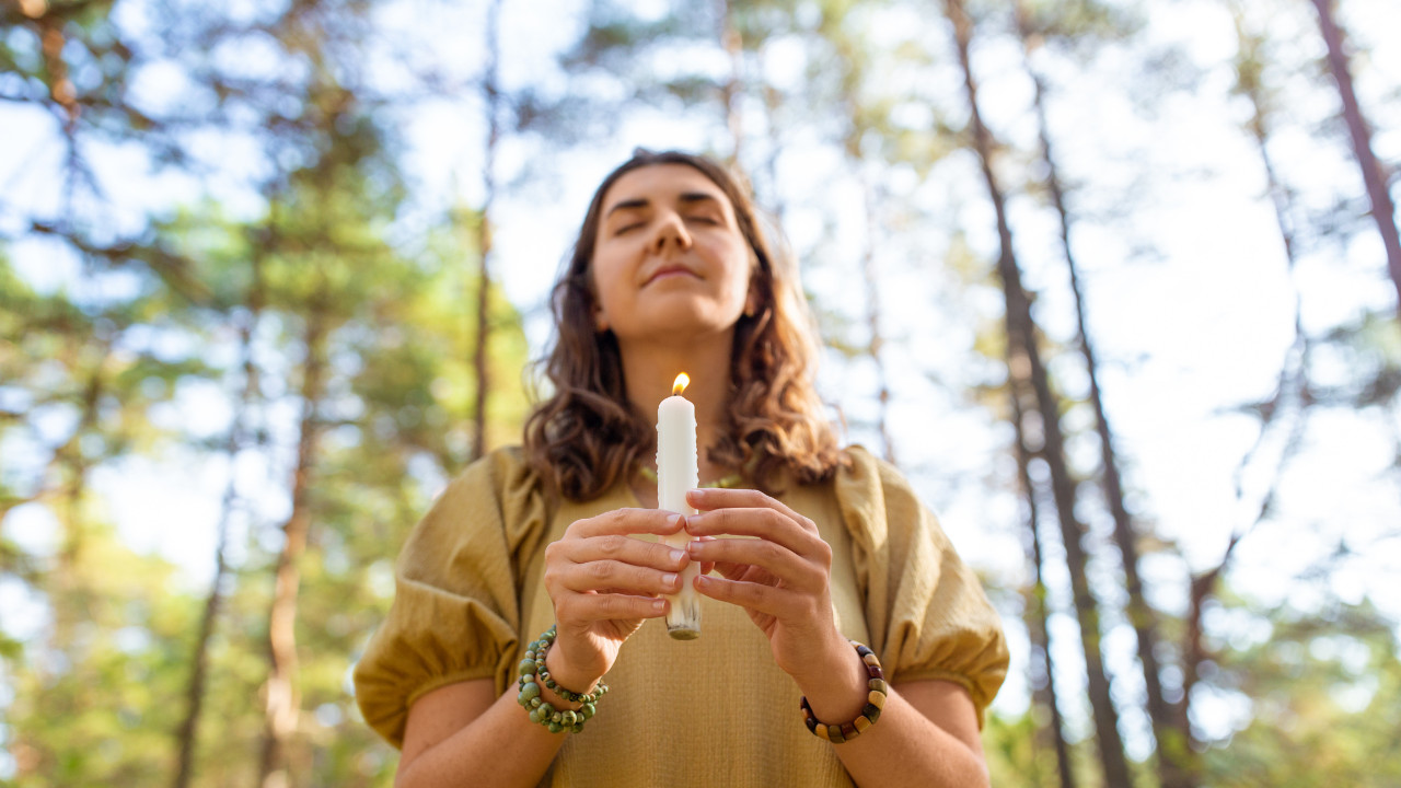 Kobieta z zamkniętymi oczami trzyma zapaloną białą świecę w lesie, skupiona i spokojna, wykonuje rytuał ochronny w otoczeniu natury.