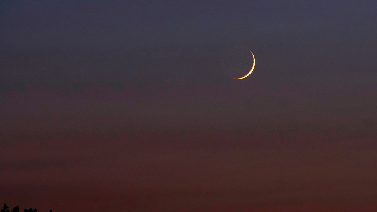 Cienki sierp Księżyca na tle ciemnego, ciepłego nieba o odcieniach czerwieni i fioletu, tuż nad linią horyzontu.