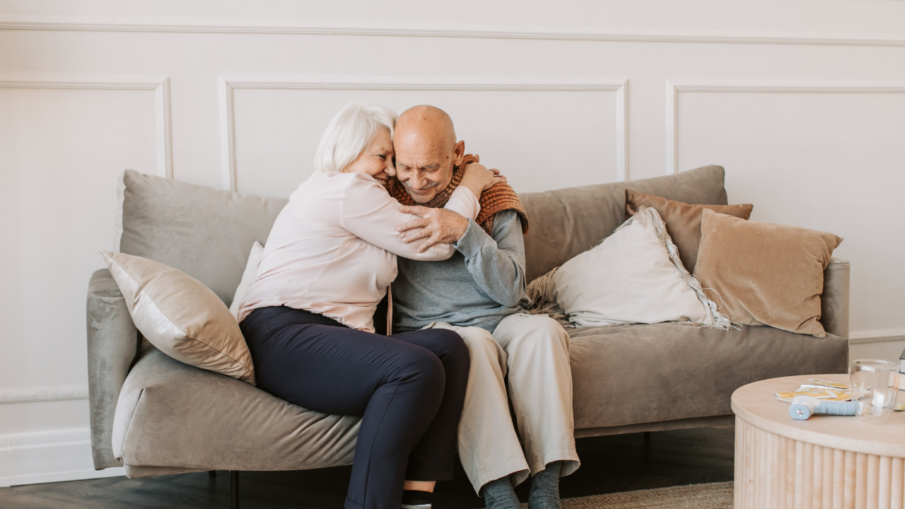 Starsza para siedzi na kanapie w jasnym salonie i czule się przytula, trzymając się za ręce. W tle widoczne są poduszki i stolik, a atmosfera jest spokojna i pełna bliskości.
