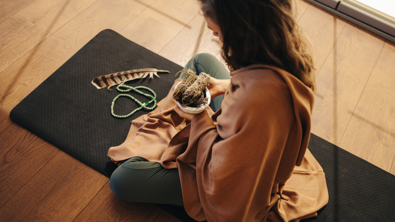 Kobieta siedzi na macie do jogi, trzyma w dłoniach suszone zioła do oczyszczania energetycznego, obok leżą pióro i koraliki