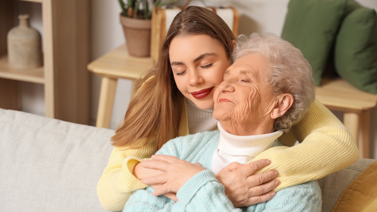 Młoda kobieta obejmuje starszą kobietę od tyłu na kanapie, obie mają zamknięte oczy i delikatnie się uśmiechają. Wnętrze jest ciepłe i przytulne, z rośliną i jasnymi meblami w tle.