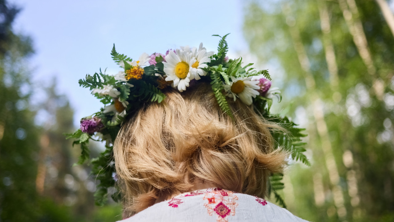 Osoba z jasnymi włosami, widziana od tyłu, ma na głowie wianek z rumianków, paproci i polnych kwiatów. Zdjęcie wykonano w zielonym lesie w słoneczny dzień, co przywołuje atmosferę tradycji związanych z Nocą Kupały i bliskością natury.