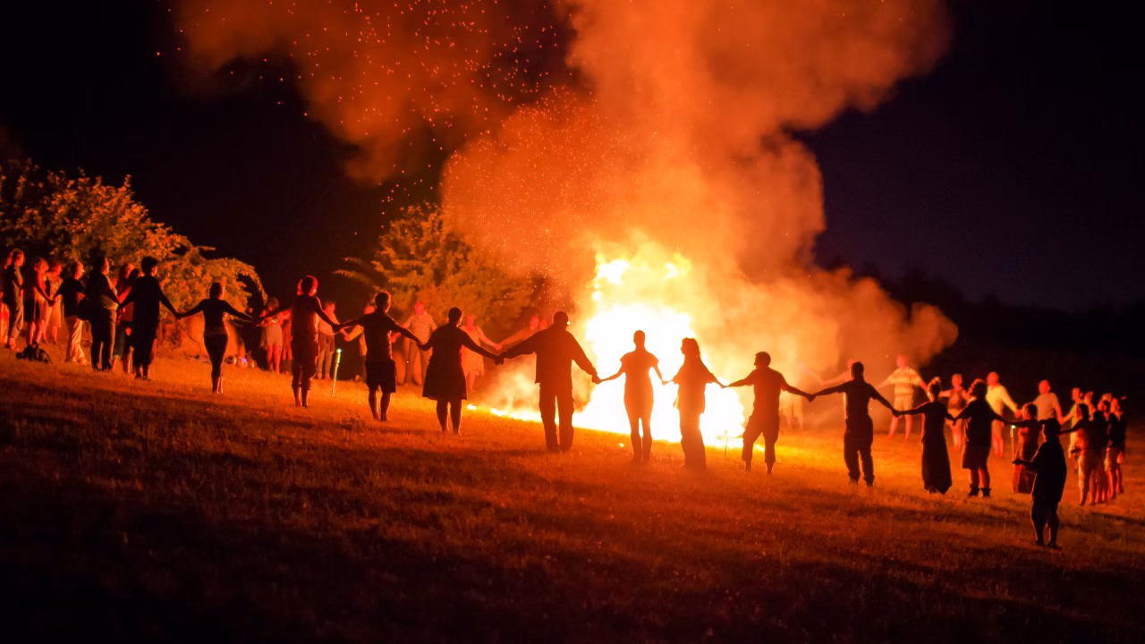 Grupa ludzi trzymających się za ręce wokół wielkiego ogniska podczas nocnego obrzędu. Płomienie i unoszące się iskry nawiązują do rytuałów na Dziady i słowiańskich tradycji.